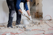 Close Up Shot Of The Legs Of A Man In A Protective Suit With A Shovel In Hand Next To A Bucket Of Debris Left Over From A House Renovation.