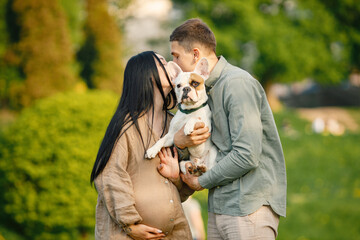 Pregnant woman and her husband standing in a park and holding little french bulldog on a hands