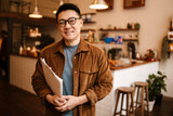 Fototapeta Na sufit - Adult asian man smiling and holding paper documents in cafe indoors