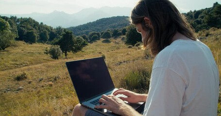 Medium shot of male freelance worker typing text message or notes on computer keyboard. Traveler on summertime journey using technology. Guy sitting on beautiful landscape with notebook.