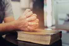 Close Up Of A Man Hands Praying On The Open Holy Bible On A Table Indoor With The Windows Light Lay Warm Tone . Christian Faith And Trust Concept With Copy Space. Christian Devotional Background