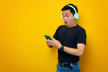 Shocked young Asian man in casual t-shirt in wireless headphones using mobile phone, checking new mobile music app isolated on yellow background. People emotions lifestyle concept