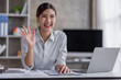 Happy positive young Asian woman enjoying online communication at home, Female using wifi while video conferencing with friend, sitting in front of open laptop, smiling and waving hand, saying hi 