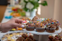 Female Hand Picking One Of Assorted Cupcakes With Sprinkles From White Plate With Blur Background