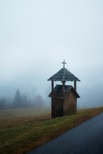 Vertical Shot Of A Small Cabin With A Cross In A Foggy Field Near The Road