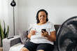 © kleberpicui - Elderly woman sitting on sofa in living room cooling off with floor fan trying to relieve heat of hot summer weather. Elderly woman drinking a glass of water on a hot day.