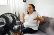 © kleberpicui - Elderly woman sitting on sofa in living room cooling off with floor fan trying to relieve heat of hot summer weather. Elderly woman drinking a glass of water on a hot day.
