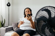© kleberpicui - Elderly woman sitting on sofa in living room cooling off with floor fan trying to relieve heat of hot summer weather. Elderly woman drinking a glass of water on a hot day.