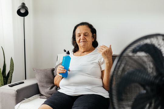 elderly woman sitting on sofa in living room cooling off with floor fan trying to relieve heat of ho