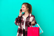 © luismolinero - Young English woman isolated on blue background holding shopping bags and calling a friend with her cell phone