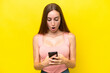 © luismolinero - Young caucasian woman isolated on yellow background looking at the camera while using the mobile with surprised expression
