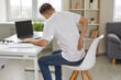 © Studio Romantic - Man experiences severe back pain caused by prolonged and incorrect sitting at workplace. Young man sitting with his back to camera holds his lower back, feeling exhausted from sedentary lifestyle.