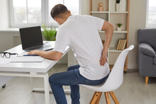 Man Experiences Severe Back Pain Caused By Prolonged And Incorrect Sitting At Workplace. Young Man Sitting With His Back To Camera Holds His Lower Back, Feeling Exhausted From Sedentary Lifestyle.
