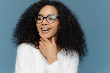 Wall Mural - Headshot of pleasant looking dark skinned young woman holds chin, looks away, has curly hair, broad smile, being in good mood, isolated on blue background. People, joy and happiness concept.