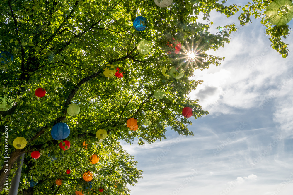 Stock-Foto „viele bunte runde Lampions hängen zwischen den Blättern eines Baumen und die Sonnenstrahlen leuchte durch die Zweige“