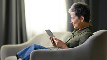 Happy Middle Aged Woman Relaxing On Couch, Using Tablet Computer, Reading News From Internet. Senior Woman Studying Online. Profile Of Middle Aged Woman In Chair With Tablet On Day Light Background