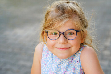 Wall Mural - Portrait of a cute preschool girl with eye glasses outdoors. Happy funny child wearing blue glasses, loooking at the camera. Sunny summer day in the city.