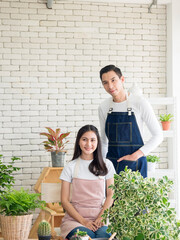 gardener young Asian man woman two person stand and sit chair smiling looking hand holding help decorate the tree leaf green in calm work shop home plant white wall. hobby job happy and care concept