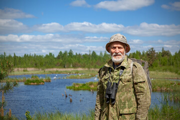 A man with a beard in camouflage clothes, with binoculars, an explorer, a tourist. Stands against the background of a forest lake, forest and blue sky with clouds.