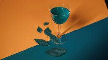 Blue Alcoholic Cocktail On Colorful Surface With Green Leaves. Stock Footage. Close Up Of A Wooman Hand Putting A Glass With Unusual Beverage On The Orange And Blue Fabric With Artificial Leaves.