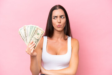 Young caucasian woman taking a lot of money isolated on pink background with sad expression