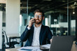 © Prostock-studio - Happy handsome businessman working on laptop and having phone conversation, sitting in office and smiling