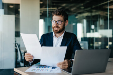 Happy businessman reading papers and business reports, rejoicing in the affairs of company, sitting in modern office