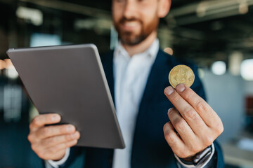 Poster - Confident businessman holding golden bitcoin and working with charts on digital tablet, standing in office, crop