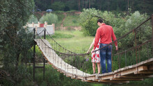 Young Lovers On A Rope Bridge Across The River. A Couple Of Hikers Traversing A Swing Bridge. Clip. Young Couple In Love Walking On Suspended Wooden Bridge Over A Lake In The Woods.