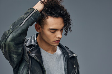 Adorable stylish tanned curly man leather jacket touches hair looks aside posing isolated on gray studio background. Cool fashion offer. Huge Seasonal Sale New Collection concept. Copy space for ad