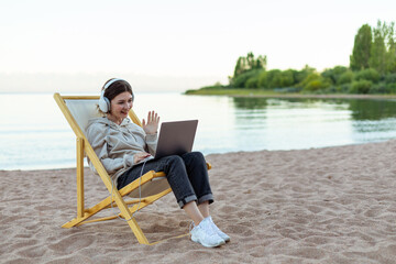 Woman in headphones video chatting by laptop, saying hello sitting on a beach