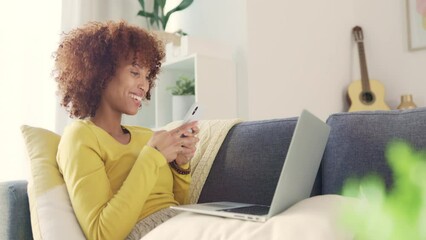 Wall Mural - Cheerful female chatting to her friends on social media, browsing online and watching funny internet memes while relaxing on a sofa. Young woman smiling and laughing while texting on a phone at home.