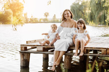 Mother Day. Happy Young Mom With Little Girls Sitting On Wooden Bridge Near Lake, Pond On Summer. Family Enjoying Life Together. Mother And Two Child Daughter Smiling While Spending Free Time Outdoors