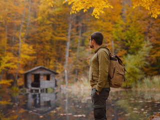 Young handsome man posing in autumn forest near lake. young hipster guy with backpack , traveller standing in woods, Hiking, Forest, Journey, active healthy lifestyle, adventure, vacation concept.