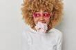 Horizontal shot of curly haired young woman has mouth full of cotton candy looks aside wears pink sunglasses and jumper isolated over white background. Female model eats appetizing sweet dessert