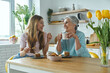 © gstockstudio - Senior mother and her adult daughter enjoying hot drinks and sweet food at the kitchen