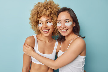 Wall Mural - Horizontal shot of mixed race young women embrace each other have friendly relationships apply beauty patches under eyes to remove puffiness stand against blue background. Skin care concept.