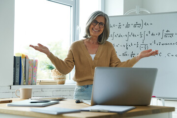 Cheerful senior woman gesturing while standing near the whiteboard at classroom