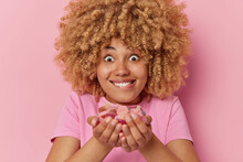 Surprised Curly Haired Young Woman Holds Jelly Candies In Hands Bites Lips Feels Temptation To Sugary Food Dressed In Casual T Shirt Isolated Over Pink Background. Unhealthy Nutrition Concept