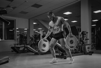 A bodybuilder in shorts is walking through the gym with two rubber plates. A shirtless sporty guy is training alone.