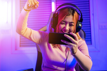 Winning. Happy Gamer playing video game online with smartphone with neon lights she raises hands to wins celebrating, Asian woman wearing gaming headphones feeling excited at home, E-Sport concept