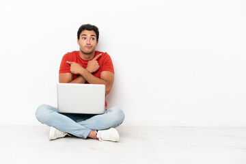 Wall Mural - Caucasian handsome man with a laptop sitting on the floor pointing to the laterals having doubts