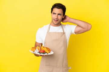 Sticker - Restaurant waiter holding waffles over isolated yellow background having doubts