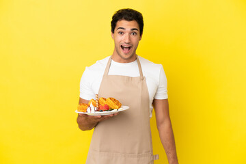 Wall Mural - Restaurant waiter holding waffles over isolated yellow background with surprise facial expression