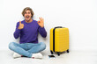 © luismolinero - Young blonde man with suitcase sitting on the floor showing ok sign and thumb up gesture
