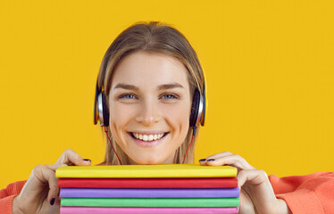 Wall Mural - Close up shot of pretty student with happy face wearing headphones holding stack of modern literature, smiling and looking at camera. Beautiful teen girl enjoying collection of good online audio books