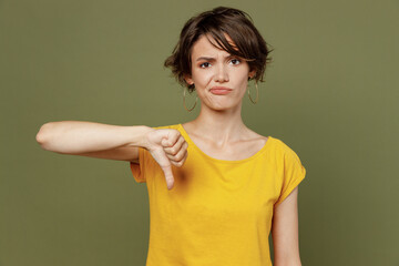 Wall Mural - Young sad displeased dissatisfied unhappy woman she 20s wear yellow t-shirt showing thumb down dislike gesture isolated on plain olive green khaki background studio portrait. People lifestyle concept.