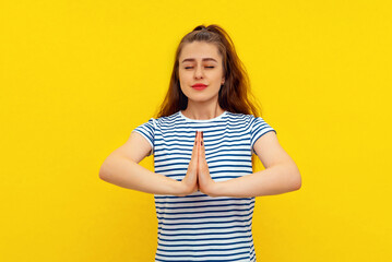 Wall Mural - Calm beautiful young woman keeps palms pressed together, showing pray, namaste gesture, express gratitude, thank you sign, standing in white-blue striped t shirt over yellow background