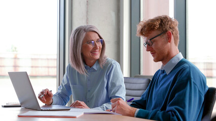 Senior grey-haired mature female teacher, executive or mentor helping young man student, new employee, teaching intern, explaining online job using laptop computer, talking, having teamwork in office.