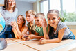 © Iryna - Cute enthusiastic little girl smiling, looking at camera and raising hand with teacher in class showing students a globe on background.